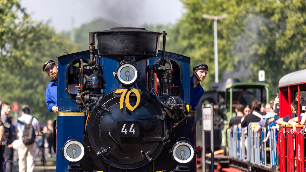 Zahlreiche Menschen stehen beim 70. Geburtstag der Cottbuser Parkeisenbahn an einem Bahnsteig, an dem eine Dampflok einfährt. Die Schmalspurbahn wird auch heute noch mit Hilfe vieler Kinder und Jugendlichen betrieben. 