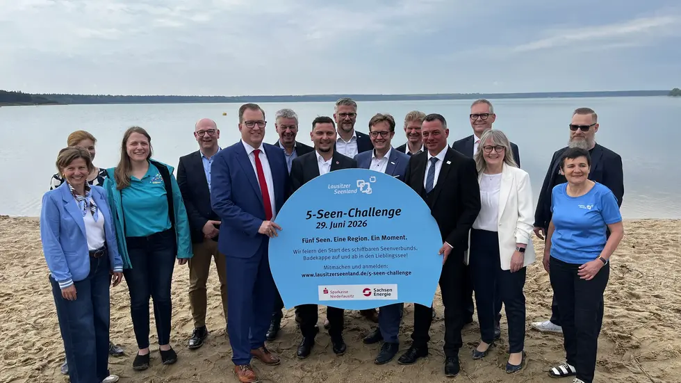 Landräte, Bürgermeister, Zweckverbände, Tourismusverband und Sponsoren werben am Badestrand des Partwitzer Sees für die 5‑Seen‑Challenge am 29. Juni 2026. Mit einem symbolischen Werbeschild in Form einer Badekappe machen sie auf die Aktion zur Eröffnung des schiffbaren Seenverbundes aufmerksam. Auf dem Foto v.l.n.r.: Constance Jacob, Sponsoring SachsenEnergie AG, Kathleen Schäfer, Unternehmenskommunikation LMBV (hinten), Alexandra Brechel, Marketing & Kommunikation LebensRäume Hoyerswerda eG, Stefan Reiter, Amtsdirektor Amt Altdöbern (hinten), Alexander Erbert, künftiger Landrat Landkreis Oberspreewald-Lausitz, Detlev Wurzler, Verbandsvorsteher Zweckverband Lausitzer Seenland Brandenburg (hinten), André Lehnick, Bürgermeister Stadt Großräschen, Daniel Just, Geschäftsführer Zweckverband Lausitzer Seenland Sachsen (hinten), Andreas Pfeiffer, Bürgermeister Stadt Senftenberg, Wolfram Korr, Geschäftsführer Brandenburgischen Sommerkonzerte/ Lausitziade (hinten), Udo Witschas, Landrat Landkreis Bautzen, Marcus Starick, Vorstandsvorsitzender Sparkasse Niederlausitz (hinten), Antje Gasterstädt, Bürgermeisterin Gemeinde Elsterheide, Kathrin Winkler, Geschäftsführerin Tourismusverband Lausitzer Seenland e.V., Tobias Dorn, Geschäftsführer KWG mbH im Lausitzer Seenland (hinten) 